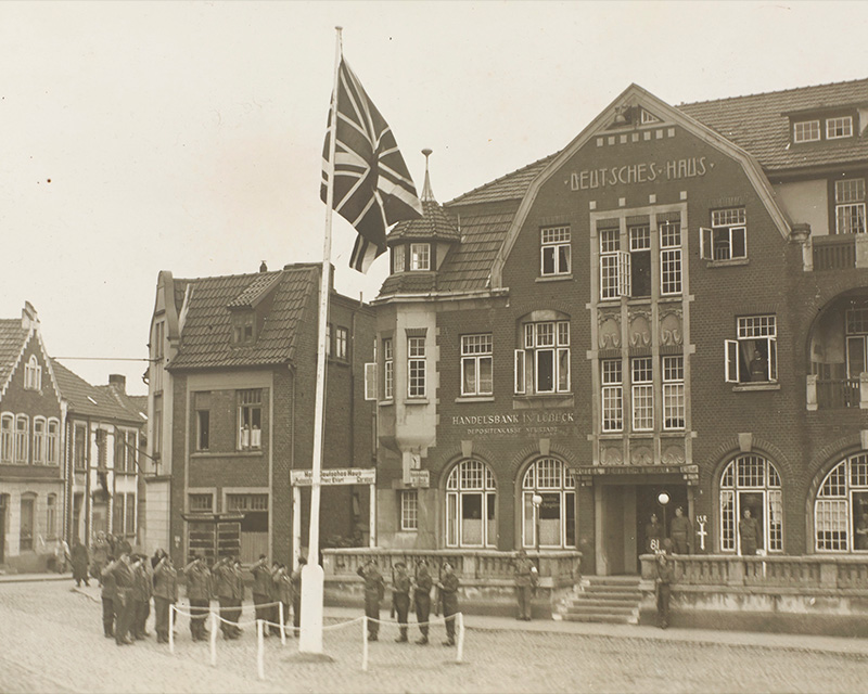 A guard and German prisoners salute the British flag, Oldenburg, Germany, May 1945