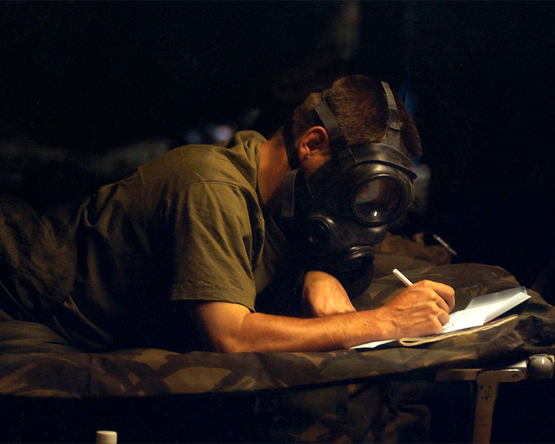 A soldier, wearing his gas mask during a chemical weapon alert, writing a letter home, northern Kuwaiti desert, 2003